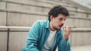 Clouseup, kot gömlekli genç sakallı adam yüksek basamaklarda oturuyor ve cep telefonu kullanıyor. Adam akıllı telefona sesli mesaj gönderiyor.