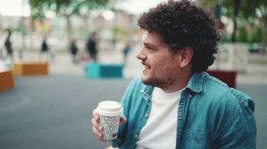 Kot gömlekli sakallı genç adamın modern şehir arka planında oturup kahve içtiği yakın plan portresi. Kahve içen adamın profili.