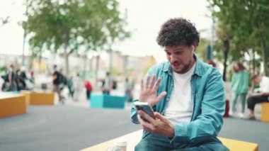 Kot tişörtlü, kablosuz kulaklıklı genç adam işlek sokak modern şehir arka planında cep telefonuyla video görüşmesi yapıyor..