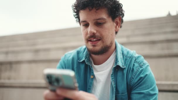 年轻大胡子男子穿着牛仔衬衫坐在高高的台阶上 使用手机的云彩肖像 男人向智能手机发送语音讯息 — 图库视频影像
