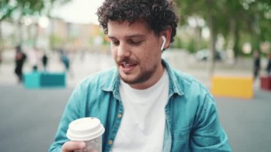 Kot gömlekli, kablosuz kulaklıklı ve modern şehir arka planında kahve içen genç sakallı adamın yakın plan portresi. Kahve içen adamın profili.