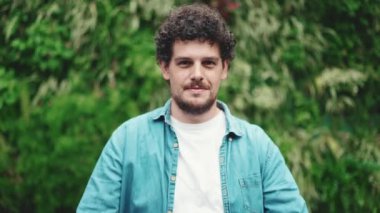 Yakın plan, kot gömlekli genç sakallı adam elini uzatıyor ve modern şehrin arka planında kameraya gülümsüyor.