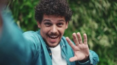 Genç sakallı adamın akış yaptığı yakın plan, yeşil yapraklı video. Adam akıllı telefon kamerasına bakarken elini sallıyor.