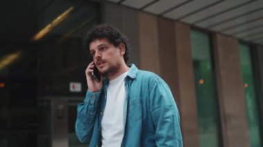 Yakın plan, kot gömlekli genç sakallı adam cep telefonuyla konuşuyor ve modern binalarda yürüyor.