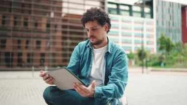 Yakın plan, kot gömlekli genç sakallı adam dışarıda tablet kullanıyor modern binalar.