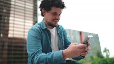 Clouseup, kot gömlekli genç sakallı adam sokakta oturuyor ve cep telefonunu kullanıyor modern binalarda.