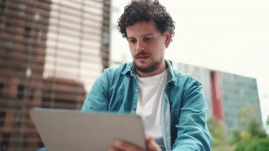 Yakın plan, kot gömlekli genç sakallı adam dışarıda tablet kullanıyor modern binalar.