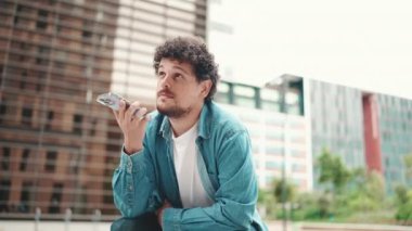 Clouseup, kot gömlekli genç sakallı adam oturuyor ve cep telefonu kullanıyor. Adam modern şehir arka planındaki akıllı telefona sesli mesaj gönderiyor.