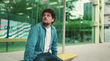 Kot pantolon giyen genç sakallı adam otobüs durağında oturur şehir taşıma saatlerini öğrenir ve modern şehir arka planında otobüs beklerken başka yere bakar.