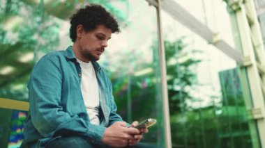 Yakın plan, kot gömlekli genç sakallı adam otobüs durağında otobüs bekliyor ve modern şehir arka planında cep telefonu kullanıyor.