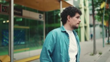 Yakın plan, kot gömlekli genç sakallı adam otobüs durağında duruyor ve modern şehir arka planında şehir ulaşımını bekliyor.