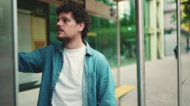 Yakın plan, kot gömlekli genç sakallı adam otobüs durağında duruyor ve modern şehir arkaplanındaki zaman çizelgesine bakıyor.