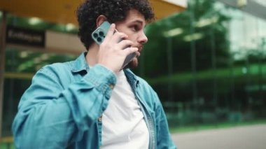 Yakın plan, kot gömlekli genç sakallı adam otobüs durağında duruyor otobüs bekliyor ve modern şehir arka planında cep telefonuyla konuşuyor.