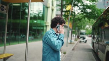 Yakın plan, kot gömlekli genç sakallı adam otobüs durağında duruyor otobüs bekliyor ve modern şehir arka planında cep telefonuyla konuşuyor.