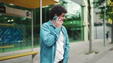 Yakın plan, kot gömlekli genç sakallı adam otobüs durağında duruyor otobüs bekliyor ve modern şehir arka planında cep telefonuyla konuşuyor.