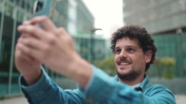 Yakından, elinde cep telefonu olan kot gömlekli genç bir adam. Modern şehir arka planında akıllı telefondan modern binaları çekiyor.