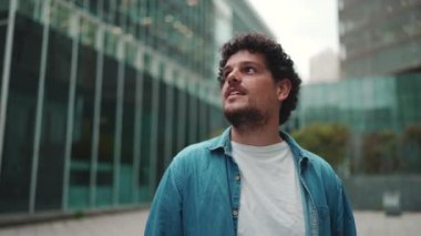 Yakın plan, kot gömlekli adam sokakta yürüyor, duruyor ve modern binaların üst katlarına bakıyor.