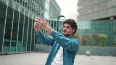 Elinde cep telefonu olan kot gömlekli genç bir adam. Adam şehrin arka planındaki akıllı telefondan modern binaları çekiyor.