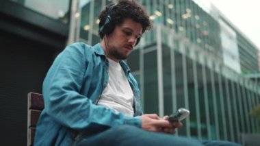 Kot gömlekli, kulaklıklı, bankta oturan, müzik dinleyen ve modern binaların arka planında akıllı telefondan yazı yazan genç sakallı adamın yakın çekimi.