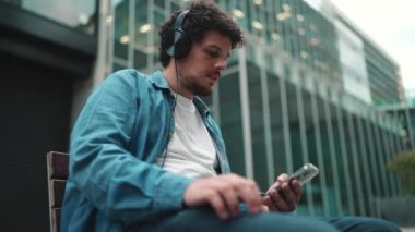 Kot gömlekli, kulaklıklı, bankta oturan, müzik dinleyen ve modern binalarda cep telefonu kullanan genç sakallı adamın yakın çekimi.