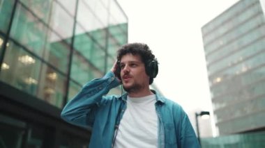 Kot pantolon giyen, kulaklık takmış, modern şehir manzarası arka planında müzik dinleyen genç sakallı adamın yakın çekimi.