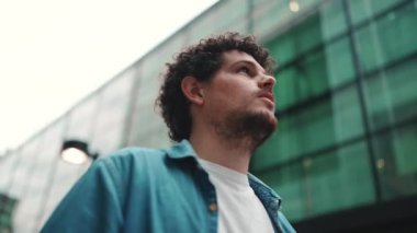 Yakın plan, kot gömlekli adam sokakta yürüyor, duruyor ve modern binaların üst katlarına bakıyor.