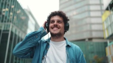 Kot pantolon giyen, kulaklık takmış, modern şehir manzarası arka planında müzik dinleyen genç sakallı adamın yakın çekimi.