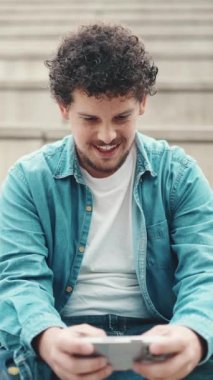 Yüksek basamaklarda oturan ve cep telefonu kullanan kot gömlekli gülümseyen sakallı genç adam. Akıllı telefonda oynayan adam.