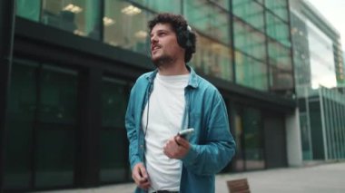 Kot gömlekli duygusal genç adam kulaklıkla sokakta yürüyor müzik dinliyor ve şarkı söylüyor, elinde cep telefonu ile modern şehir manzarası arka planında.
