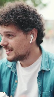 Kot tişörtlü, kablosuz kulaklıkla oturan ve modern şehir arka planında kahve içen genç sakallı adamın yakın plan portresi. Kahve içen adamın profili.