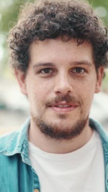 Kot tişörtlü genç sakallı bir adamın modern şehir manzarası arka planında poz verirken ve gülümserken ön yakın plan portresi..