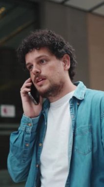 Yakın plan, kot gömlekli genç adam cep telefonuyla konuşuyor ve modern binaların arka planında yürüyor.