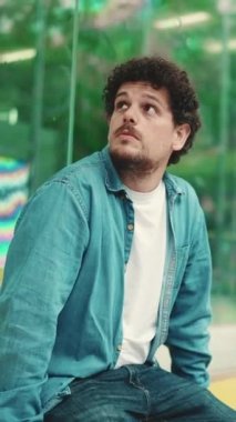 Kot gömlekli genç sakallı adam otobüs durağında oturmuş şehir taşıma programlarını çalışıyor ve modern şehir arka planında otobüs beklerken başka tarafa bakıyor.