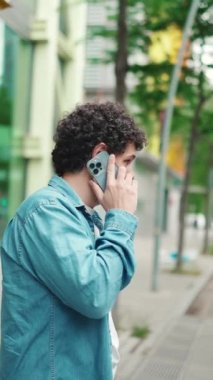 Yakından, kot gömlekli genç sakallı adam otobüs durağında duruyor otobüs bekliyor ve modern şehir arka planında cep telefonuyla konuşuyor.