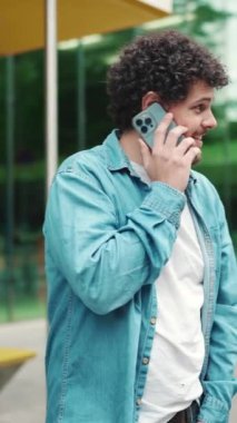 Yakından, kot gömlekli genç sakallı adam otobüs durağında duruyor otobüs bekliyor ve modern şehir arka planında cep telefonuyla konuşuyor.