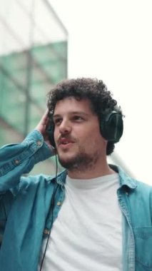 Kot gömlekli, kulaklık takmış, modern şehir manzarası arka planında müzik dinleyen genç sakallı bir adamın yakın çekimi.