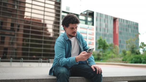 身穿斜纹棉布衬衫 留着胡子的年轻人坐在街上 用手机在现代建筑的背景下 — 图库视频影像