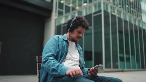 Joven Barbudo Con Camisa Mezclilla Sentado Auriculares Banco Escuchando Música — Vídeos de Stock