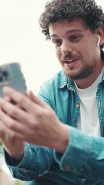 Kot gömlekli, kablosuz kulaklıklı genç sakallı adam işlek sokak modern şehir arka planında cep telefonundan video görüşmesi yapıyor..