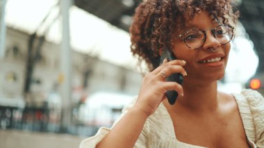 Gözlüklü genç bir kadının yakın plan portresi istasyonda oturuyor ve akıllı telefondan konuşuyor. Şehir arka planında cep telefonu kullanan olumlu kadın..