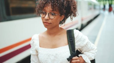 Gözlüklü, sırt çantalı genç bir kadının yakın plan portresi trenin yanındaki platformda duruyor. 