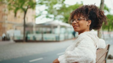 Gözlüklü genç bir kadın sırıtarak bankta oturuyor ve etrafına bakıyor. Mutlu kız bir bankta otururken etrafına bakar