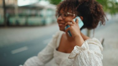 Gözlüklü genç bir kadın açık hava bankında oturmuş gülümsüyor ve akıllı telefonda konuşuyor. Şehir arka planında cep telefonu kullanan olumlu kadın..