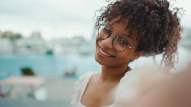 Gözlüklü genç bir kadın limanda dikiliyor ve akıllı telefondan selfie çekiyor. Şehir arka planında cep telefonu kullanan olumlu kadın..