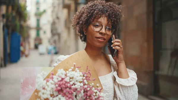 Young woman in glasses walks with a bouquet of flowers along the street of the old city and talks on a smartphone. Positive woman using mobile phone outdoors in urban background.