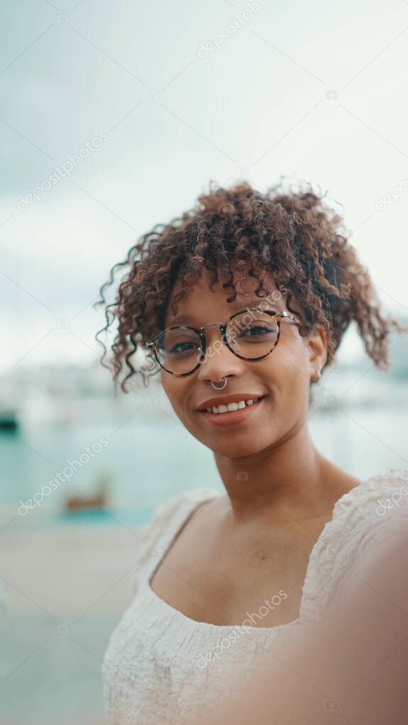 Closeup of a young woman in glasses stands in the seaport and shots selfie on smartphone. Positive woman using mobile phone outdoors in urban background.