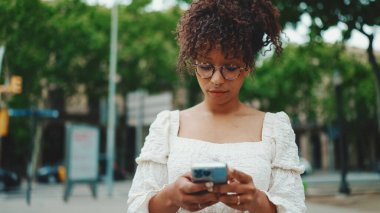 Gözlüklü genç bir kadın elinde akıllı telefonuyla ayakta duruyor ve etrafına bakıyor. Şehir arka planında cep telefonu kullanan kadın.