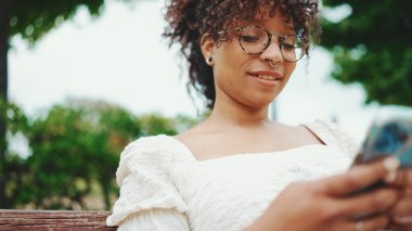 Parktaki bankta elinde akıllı bir telefonla oturan genç bir kadının yakın plan portresi. Şehir arka planında cep telefonu kullanan olumlu kadın..