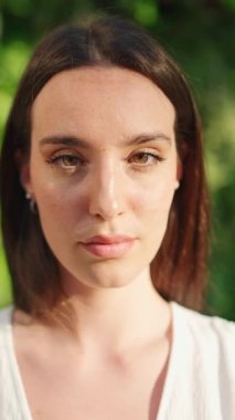 Çilli, koyu renk saçlı, beyaz bluzlu ve kot pantolonlu genç bir kadın kameraya bakıp gülümsüyor. Uzun kirpikleri olan güzel bir kız kameraya gülümsüyor.