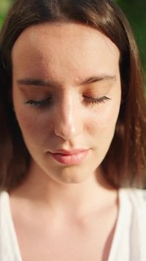 Çilli, koyu renk saçlı, beyaz bluzlu ve kot pantolonlu genç bir kadın gözlerini açıyor ve gülümsüyor. Uzun kirpikleri olan güzel bir kız kameraya gülümsüyor.